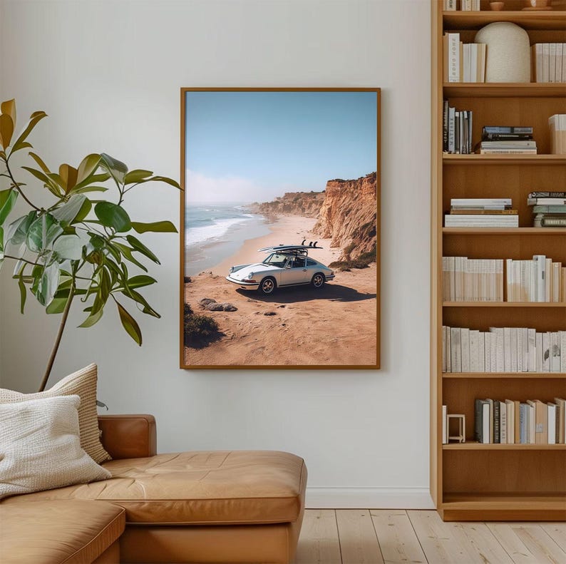 May include: A framed print featuring a vintage silver car parked on a sandy beach with surfboards on the roof. The scene includes a blue sky, ocean waves, and coastal cliffs. The print is displayed in a room with a leather couch and a bookshelf.