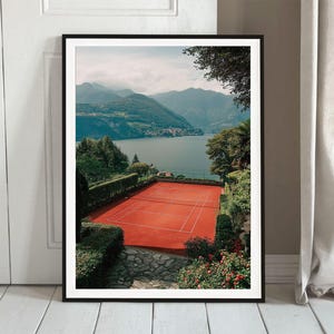 Peut inclure: Impression encadrée d'un court de tennis en terre battue rouge avec des lignes blanches, surplombant un lac et des montagnes. La scène est entourée de verdure et d'une ville au loin. Le ciel est nuageux.