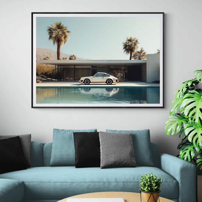 May include: Framed art print featuring a white sports car parked near a pool in front of a modern home. Palm trees and mountains are visible in the background under a clear sky. The artwork is displayed above a blue sofa with black and patterned pillows.
