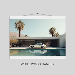 May include: A framed print featuring a white sports car parked in front of a modern home with a pool. Palm trees and mountains are in the background under a clear blue sky. The print is displayed with a white wood hanger.