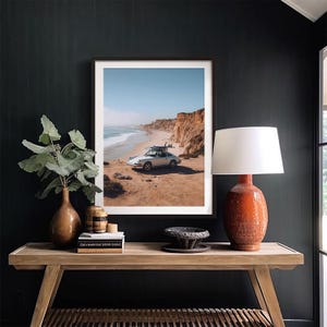 May include: Framed print of a white vintage car parked on a sandy beach with surfboards on top. The ocean and cliffs are in the background under a blue sky. The print is displayed on a dark wall.