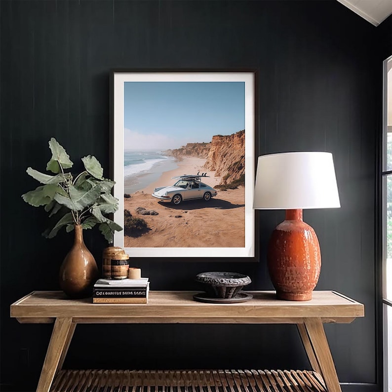 May include: Framed print of a white vintage car parked on a sandy beach with surfboards on top. The ocean and cliffs are in the background under a blue sky. The print is displayed on a dark wall.