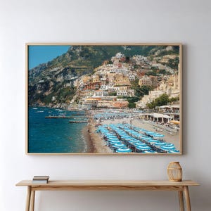 Può includere: Stampa incorniciata di una città costiera con edifici colorati su una collina che domina una spiaggia. Ombrelloni turchesi e bianchi costeggiano la riva, con barche sul mare blu. Il cielo è sereno.