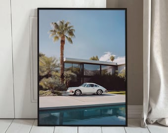 May include: A framed wall art print featuring a white vintage sports car parked on a concrete driveway in front of a modern house with large glass windows, surrounded by palm trees and a swimming pool reflecting the clear blue sky above.