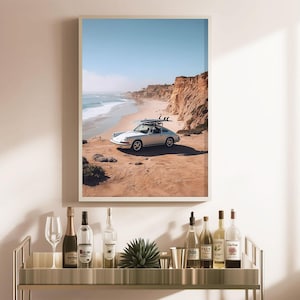May include: Framed print of a silver vintage car with surfboards on top, parked on a sandy beach. The ocean and cliffs are in the background under a clear blue sky. The print is displayed above a bar cart with bottles and glasses.