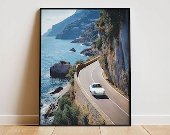 May include: A framed wall art print depicting a white vintage sports car driving along a winding coastal road. The scene features a steep cliffside, deep blue ocean waters, and a distant town nestled into the mountainside under a clear sky.