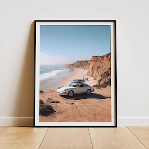 May include: Framed print of a white vintage car parked on a sandy beach with surfboards on the roof. The car is facing the ocean, with a clear blue sky and cliffs in the background. The print is in a black frame.
