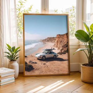 May include: A framed print of a vintage silver car with surfboards parked on a sandy beach next to a cliff. The sky is blue, and the ocean waves are visible. The frame is a light brown color.