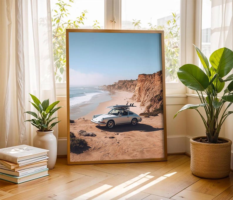 May include: A framed print of a vintage silver car with surfboards parked on a sandy beach next to a cliff. The sky is blue, and the ocean waves are visible. The frame is a light brown color.