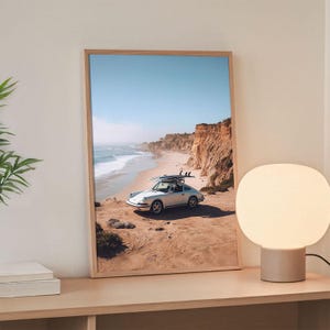 May include: Framed print of a white vintage car with surfboards on top, parked on a sandy beach. The car faces the ocean, with cliffs and a clear blue sky in the background. The print is in a light wood frame.