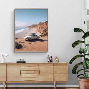 May include: A framed print of a white vintage car with surfboards on top, parked on a sandy cliff overlooking the ocean. The sky is blue, and the cliffs are a reddish-brown color.