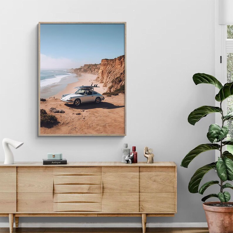 May include: A framed print of a white vintage car with surfboards on top, parked on a sandy cliff overlooking the ocean. The sky is blue, and the cliffs are a reddish-brown color.