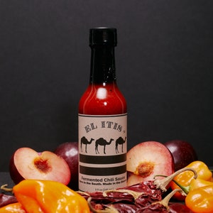 May include: A bottle of "El Itis" fermented chili sauce with a black cap and a label featuring three camel silhouettes. The bottle is surrounded by fresh plums, orange peppers, and dried red chilies. The label reads "Born in the South. Made in the Desert."