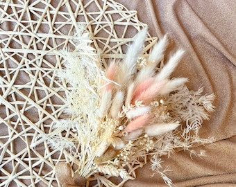 Può includere: Un bouquet di fiori secchi con accenti rosa pallido e bianco, legato con un fiocco di spago naturale. Il bouquet è appoggiato su un sottobicchiere intrecciato a forma di stella. Lo sfondo è un tessuto scintillante color beige.