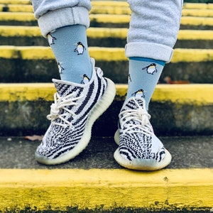 May include: A pair of white and black zebra-print sneakers with white laces. The sneakers are worn by a person wearing light blue sweatpants and blue socks with a penguin pattern.