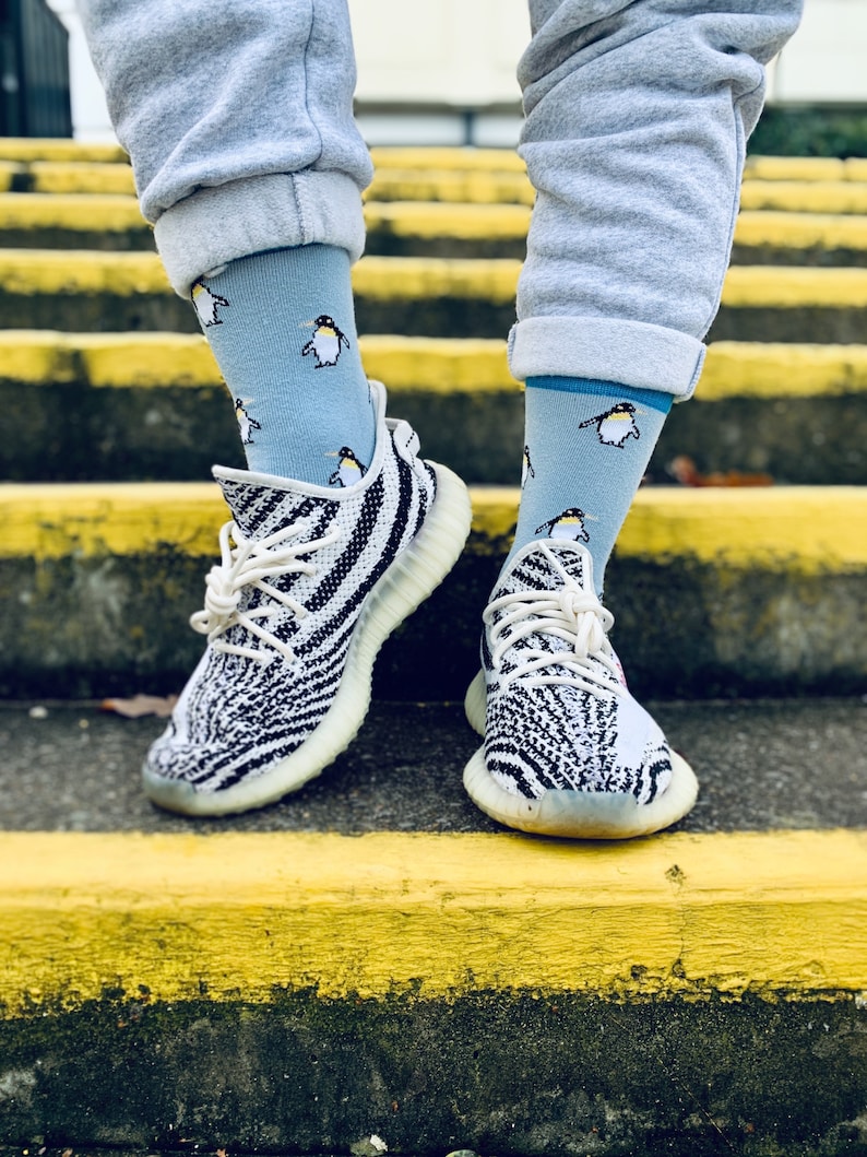 May include: A pair of white and black zebra-print sneakers with white laces. The sneakers are worn by a person wearing light blue sweatpants and blue socks with a penguin pattern.