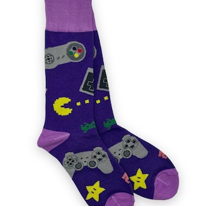 May include: Purple socks with a retro video game theme. The socks feature a gray and black video game controller, a yellow Pac-Man, a green space invader, and a yellow star.