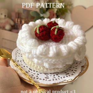 Könnte beinhalten: Ein Häkelmuster für einen weißen Kuchen mit roten und grünen Erdbeeren darauf. Der Kuchen befindet sich auf einer weißen Spitze und einem weißen Teller mit goldfarbenem Rand. Der Text "PDF PATTERN" befindet sich oben im Bild. Der Text "not a physical product <3" befindet sich unten im Bild.
