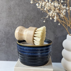 May include: A blue ceramic dish with a wooden-handled brush with natural bristles. The dish is sitting on a light wood platform.