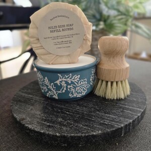 K&ouml;nnte beinhalten: Ein runder, beiger Sp&uuml;lmittel-Nachf&uuml;llriegel mit dem Text "Rooted Essentials Solid Dish Soap Refill Rounds" und einer Zutatenliste. Der Seifenriegel liegt in einer blauen Keramikschale mit wei&szlig;em Blumenmuster. Neben der Schale befindet sich eine B&uuml;rste aus Holz mit Naturborsten. Die Schale und die B&uuml;rste befinden sich auf einer runden schwarzen Steinoberfl&auml;che.