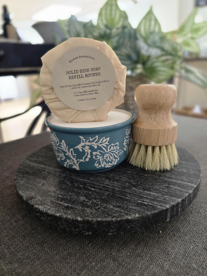 K&ouml;nnte beinhalten: Ein runder, beiger Sp&uuml;lmittel-Nachf&uuml;llriegel mit dem Text "Rooted Essentials Solid Dish Soap Refill Rounds" und einer Zutatenliste. Der Seifenriegel liegt in einer blauen Keramikschale mit wei&szlig;em Blumenmuster. Neben der Schale befindet sich eine B&uuml;rste aus Holz mit Naturborsten. Die Schale und die B&uuml;rste befinden sich auf einer runden schwarzen Steinoberfl&auml;che.