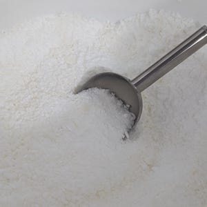 May include: A white bowl filled with a white, grainy substance, such as powdered sugar or coconut flakes. A silver scoop is partially submerged in the substance.