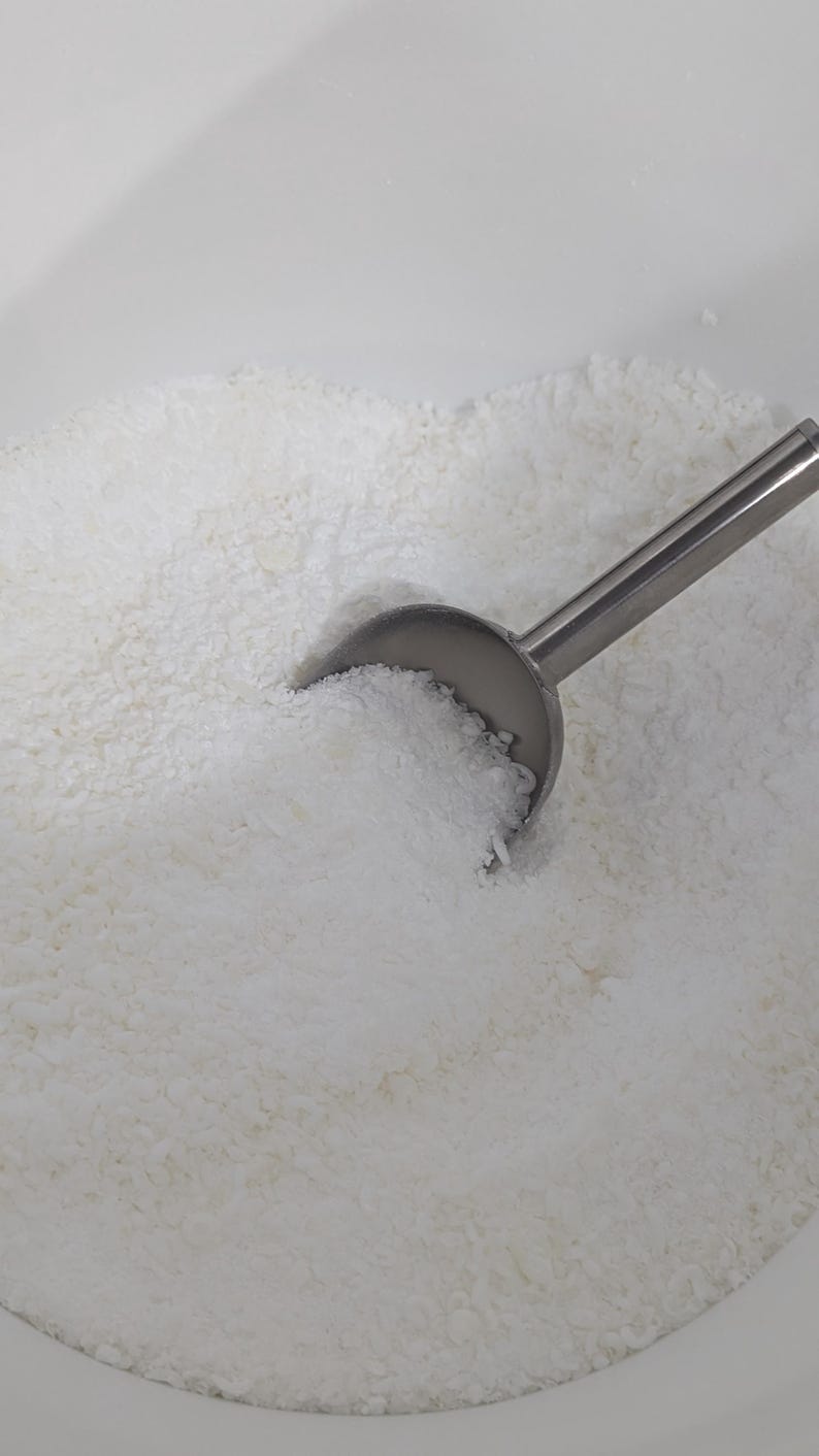 May include: A white bowl filled with a white, grainy substance, such as powdered sugar or coconut flakes. A silver scoop is partially submerged in the substance.