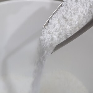 May include: A white powder is being poured from a metal scoop into a white bowl.