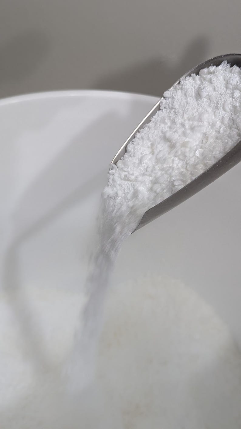May include: A white powder is being poured from a metal scoop into a white bowl.