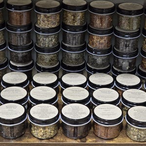 May include: A collection of small glass jars filled with various dried herbs and spices. The jars are arranged in rows on a wooden shelf. Each jar has a label with the name of the herb or spice.