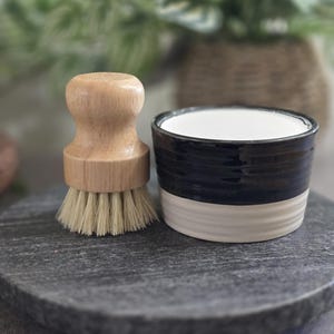 以下が含まれることがあります： 白と黒のセラミック製の皿と、天然毛のついた木製のブラシ。皿とブラシは、濃い灰色の石の表面の上に置かれています。