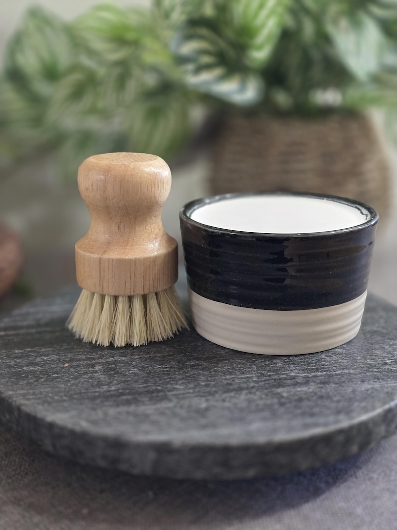 以下が含まれることがあります： 白と黒のセラミック製の皿と、天然毛のついた木製のブラシ。皿とブラシは、濃い灰色の石の表面の上に置かれています。