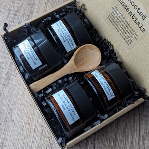 May include: A set of four small glass jars with black lids, filled with a brown substance. The jars are labeled with the names of different face masks: "Deep Cleanse", "Minerals & Antioxidants", "Rejuvenate & Hydrate", and "Soothe & Calm". The jars are arranged in a brown cardboard box with black shredded paper. A wooden spoon is included in the box.