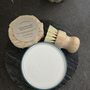 K&ouml;nnte beinhalten: Ein runder, in braunes Papier gewickelter Block fester Sp&uuml;lmittel mit dem Text "Solid Dish Soap Refill Rounds" und einer Zutatenliste. Die Seife liegt neben einer B&uuml;rste mit Naturborsten und einem wei&szlig;en Keramikgeschirr mit blauem Rand.