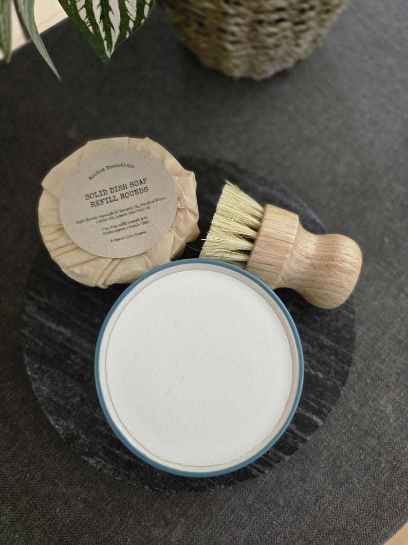 K&ouml;nnte beinhalten: Ein runder, in braunes Papier gewickelter Block fester Sp&uuml;lmittel mit dem Text "Solid Dish Soap Refill Rounds" und einer Zutatenliste. Die Seife liegt neben einer B&uuml;rste mit Naturborsten und einem wei&szlig;en Keramikgeschirr mit blauem Rand.