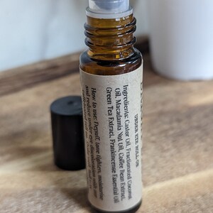 May include: A brown glass roller bottle with a silver roller ball. The bottle is labeled "UNDER EYE ROLL ON" and lists the ingredients: Castor Oil, Fractionated Coconut Oil, Macadamia Nut Oil, Coffee Bean Extract, Green Tea Extract, Frankincense Essential Oil. The label also includes instructions on how to use the product: "Depuff, tone, lighten, moisturize and reduce under eye discoloration with our caffeinated roll on. Shake well. Roll on."