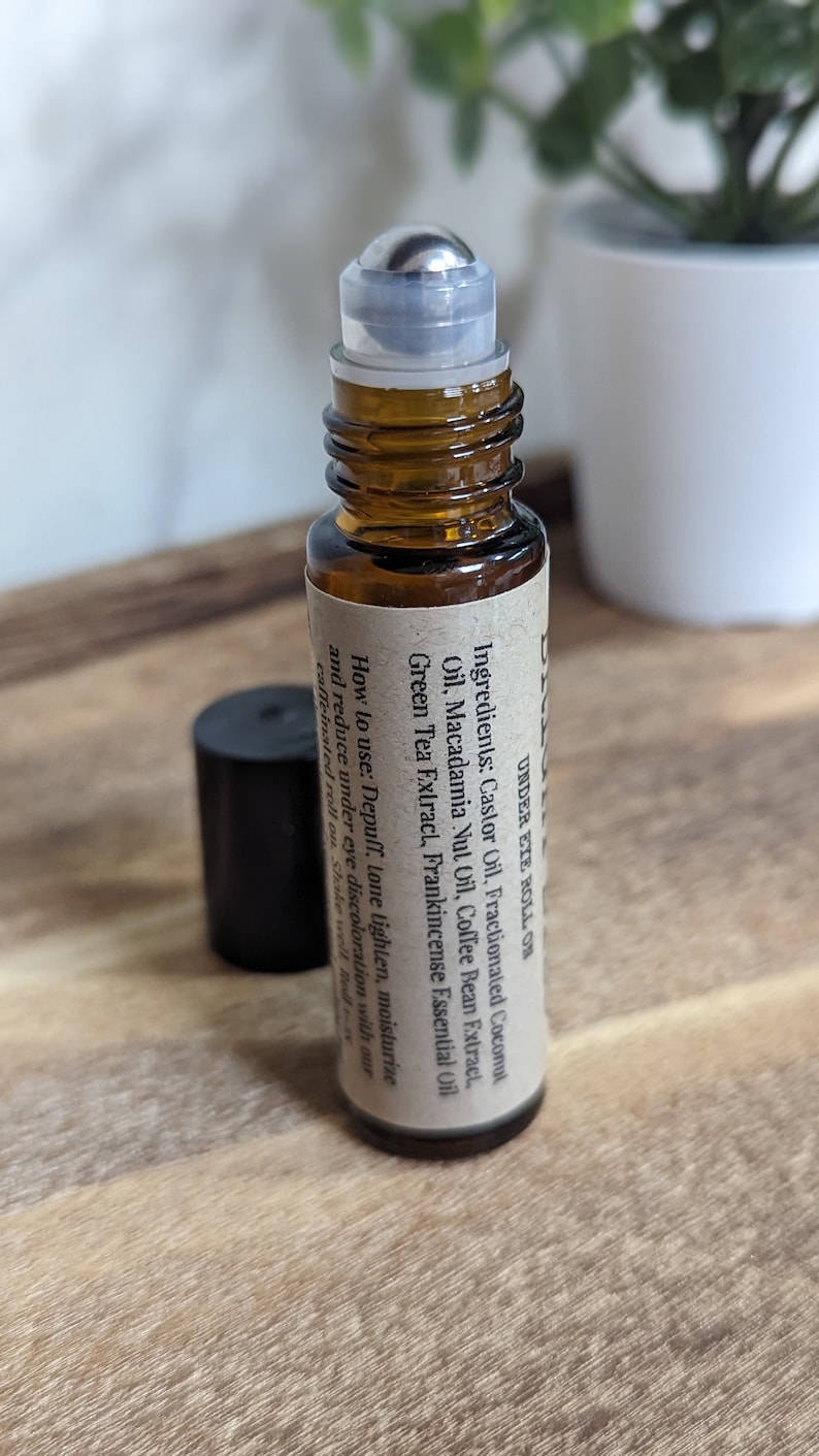 May include: A brown glass roller bottle with a silver roller ball. The bottle is labeled "UNDER EYE ROLL ON" and lists the ingredients: Castor Oil, Fractionated Coconut Oil, Macadamia Nut Oil, Coffee Bean Extract, Green Tea Extract, Frankincense Essential Oil. The label also includes instructions on how to use the product: "Depuff, tone, lighten, moisturize and reduce under eye discoloration with our caffeinated roll on. Shake well. Roll on."