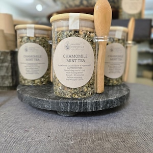 May include: A display of chamomile mint tea in clear jars with wooden lids. Each jar has a label with the text "Chamomile Mint Tea". A wooden spoon rests in front of the jars. The jars sit on a dark gray, round stone display.