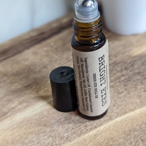 May include: A brown glass roller bottle with a silver roller ball and a brown paper label that reads "BRIGHT EYES UNDER EYE ROLL ON" and lists the ingredients: Castor Oil, Fractionated Coconut Oil, Macadamia Nut Oil, Coffee Bean Extract, Green Tea Extract, Frankincense Essential Oil.