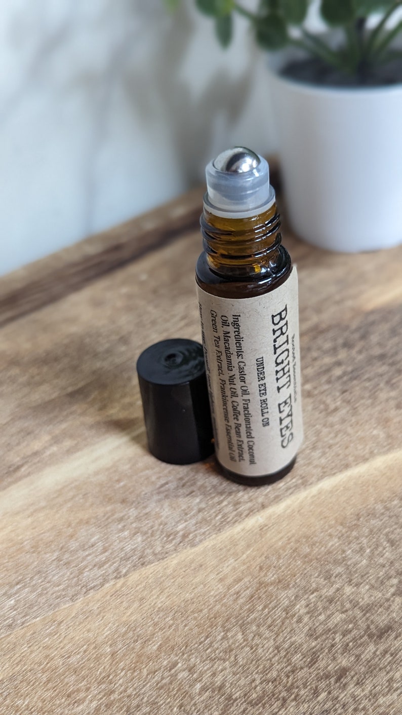 May include: A brown glass roller bottle with a silver roller ball and a brown paper label that reads "BRIGHT EYES UNDER EYE ROLL ON" and lists the ingredients: Castor Oil, Fractionated Coconut Oil, Macadamia Nut Oil, Coffee Bean Extract, Green Tea Extract, Frankincense Essential Oil.