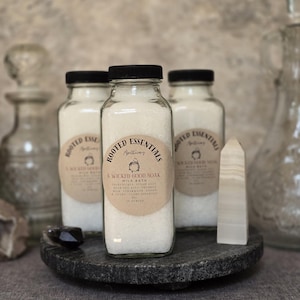 May include: Three clear glass bottles filled with white milk bath powder, each topped with a black cap and a round tan label. The label reads Rooted Essentials Apothecary, A Wicked Good Soak, Milk Bath, ingredients, and 10 ounces.