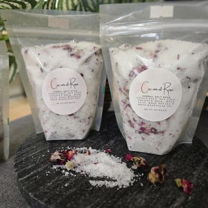 May include: Two clear pouches filled with herbal salt soak, Epsom salt, coconut milk, rose petals, and rose essential oil. The pouches have a circular label that reads "Coconut Rose". The product is displayed on a dark gray stone surface.
