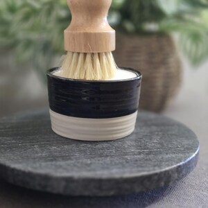 以下が含まれることがあります： 黒と白のセラミック製のブラシホルダーで、木製のブラシハンドルと白いブラシが付いています。ブラシホルダーは灰色の石の表面の上に置かれています。