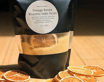May include: A black pouch with a white label that reads "Orange Dream Mineral Bath Salts" and lists the ingredients: Epsom salt, yellow clay, turmeric, essential oils of orange, tangerine & mandarin, dried oranges, orange peel. The pouch contains a yellow bath salt mixture with dried orange slices. The pouch is sitting on a wooden surface with dried orange slices.