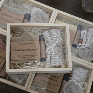 May include: Four wooden boxes with clear lids, each containing a bar of soap, a small bottle of essential oil, and a natural sponge. The soap bars are labeled with the names "Dandelion & Hemp", "Vanilla Oatmeal", "Lemongrass Patchouli", and "Rooted Essentials".