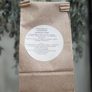 May include: A brown paper bag with a white label that reads "Rooted Essentials Shredded Laundry Soap". The label lists the ingredients as saponified coconut oil, sodium percarbonate, sodium carbonate, and essential oil. It also includes instructions for use, stating to add 1 tablespoon for small/medium loads or 2 tablespoons for large/super loads. The label also states that the soap is biodegradable, safe for septic systems, SLS free, detergent free, and eco-friendly. The bag contains 10 ounces (283.5 g) of shredded laundry soap.
