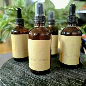 May include: Four brown glass bottles with black tops and cream-colored labels are arranged on a dark gray stone surface. The labels read "Goddess Oil" and "Goddess Hydrosol" with illustrations. The bottles are in front of green leafy plants.