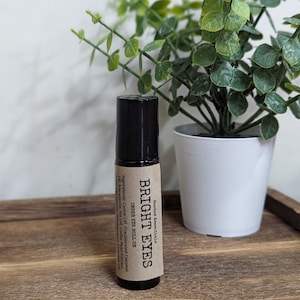 May include: A brown glass roll-on bottle with a black cap and a brown paper label that reads "Rooted Essentials BRIGHT EYES UNDER EYE ROLL ON Ingredients: Castor Oil, Fractionated Coconut Oil, Macadamia Nut Oil, Coffee Bean Extract, "