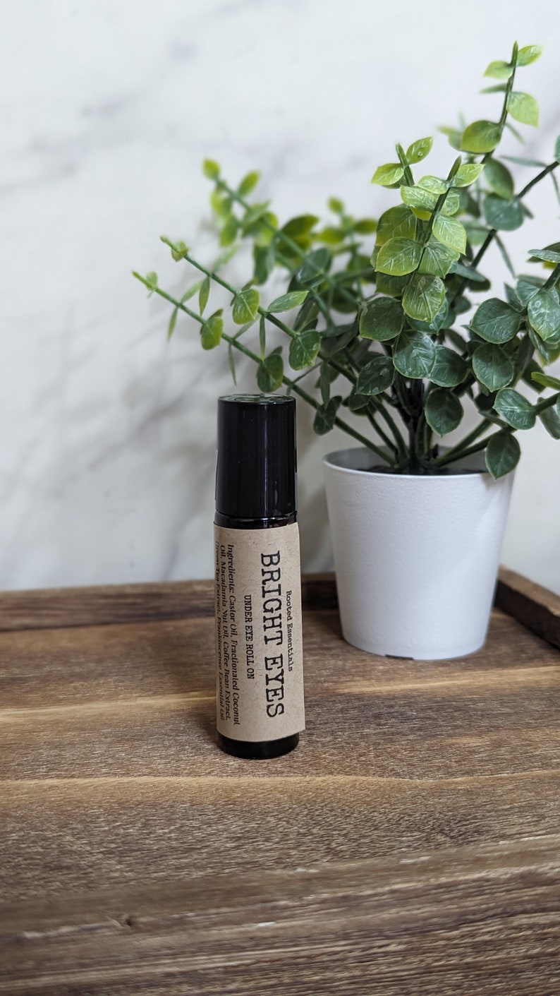May include: A brown glass roll-on bottle with a black cap and a brown paper label that reads "Rooted Essentials BRIGHT EYES UNDER EYE ROLL ON Ingredients: Castor Oil, Fractionated Coconut Oil, Macadamia Nut Oil, Coffee Bean Extract, "