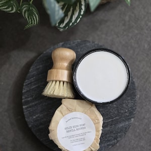 以下が含まれることがあります： 白い丸い食器用石鹸のリフィルバーで、ラベルに「Rooted Essentials Solid Dish Soap Refill Rounds」と書いてあります。石鹸は黒いスレートの表面に置かれ、木製のブラシと白いセラミックの容器があります。
